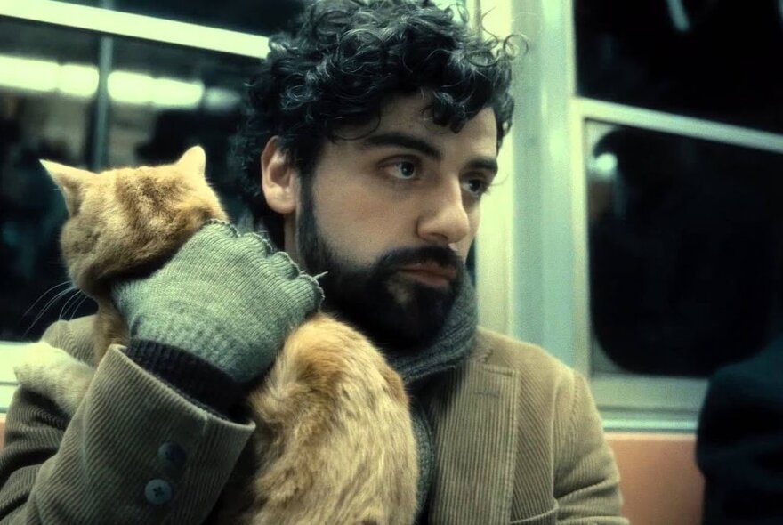 A film still showing a man wearing gloves, a coat and scarf holding a ginger cat close to his chest, on public transport, possibly a train.