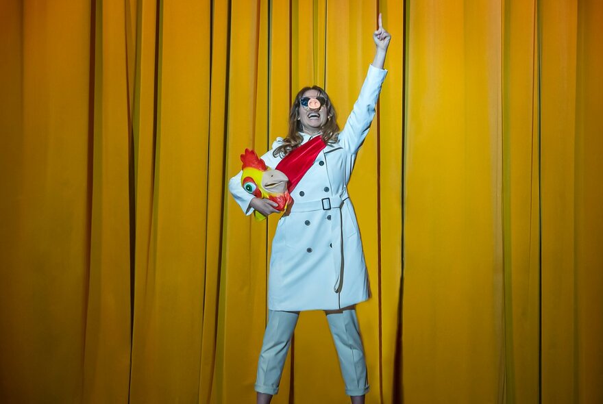 A performer wearing a white trench coat and a pig's snout nose, carrying a chicken head mask under one arm, her other arm raised above her head, standing on a stage in front of a yellow curtain.