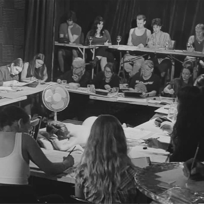 A black and white protograph of life drawing students seated at desks surrounding a life model lounging in a pose in the centre.