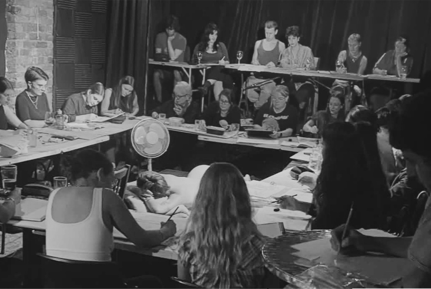 A black and white protograph of life drawing students seated at desks surrounding a life model lounging in a pose in the centre.