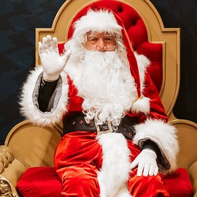 Santa sitting on his throne with one hand in the air waving. 