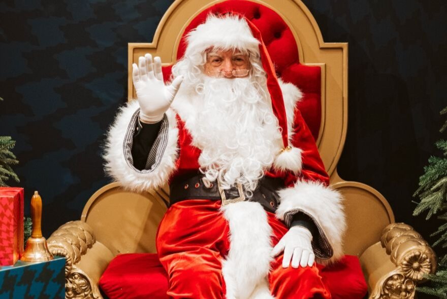 Santa sitting on his throne with one hand in the air waving. 