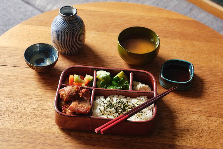 A ceramic bento box, each section filled with morsels of Japanese food, with small ceramic vessels of sauces and sake on the wooden table next to it.