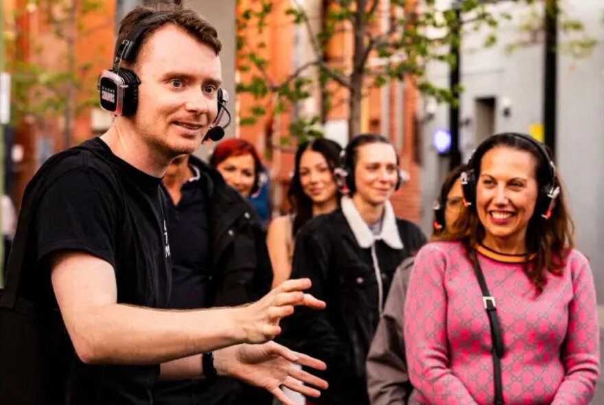 A group of people with headphones on listening to a man in black as he performs a walking comedy show.