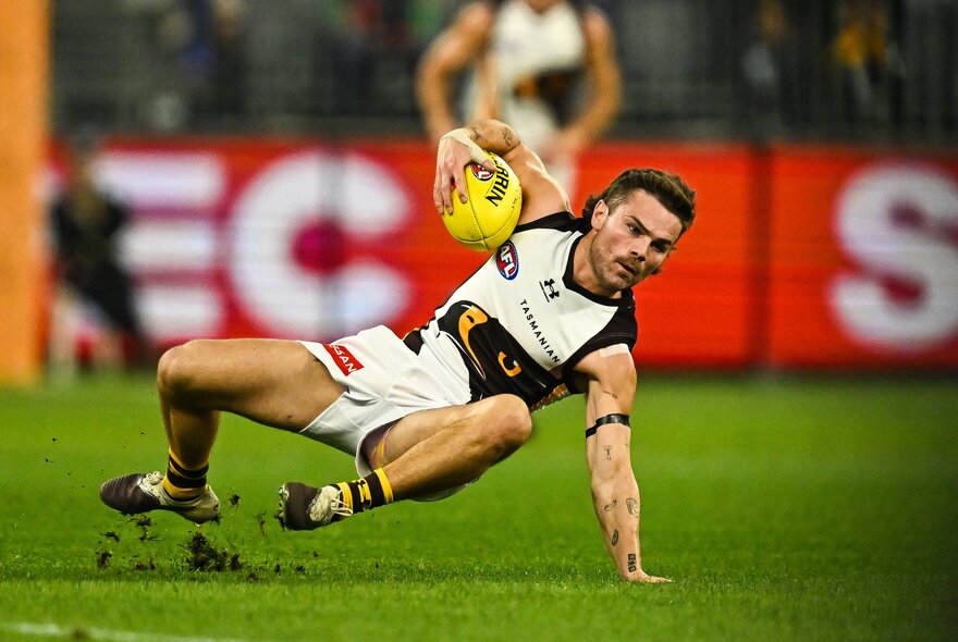 A Hawthorn AFL player holding the ball in one hand, and skidding into the field during a match.