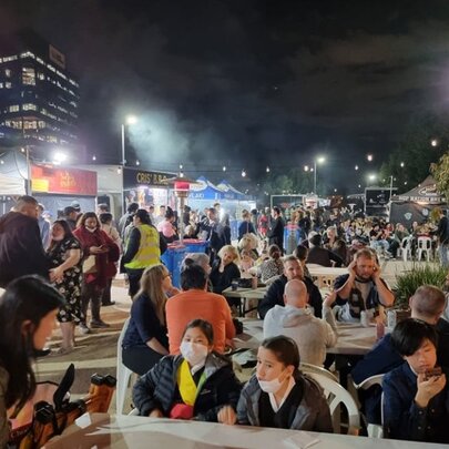 Outdoors at night, people seated in chairs at tables, some walking around, in the vicinity of the open plaza at Queen Victoria Market.