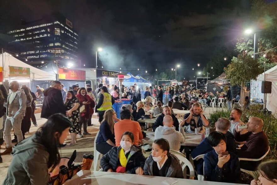Outdoors at night, people seated in chairs at tables, some walking around, in the vicinity of the open plaza at Queen Victoria Market.