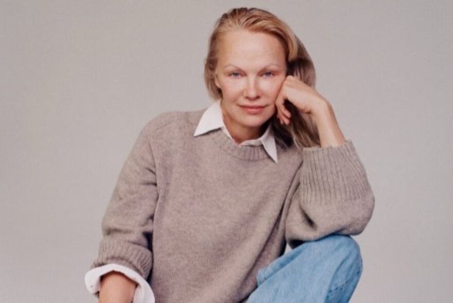 Portrait of 90s icon, Pamela Anderson, resting her face on her hand, wearing a knit jumper over a white shirt and blue jeans.