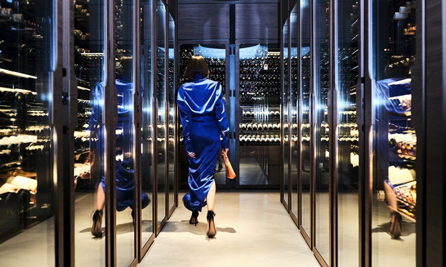 A woman in a blue silk dress walking through a restaurant hallway lined with wine bottles.