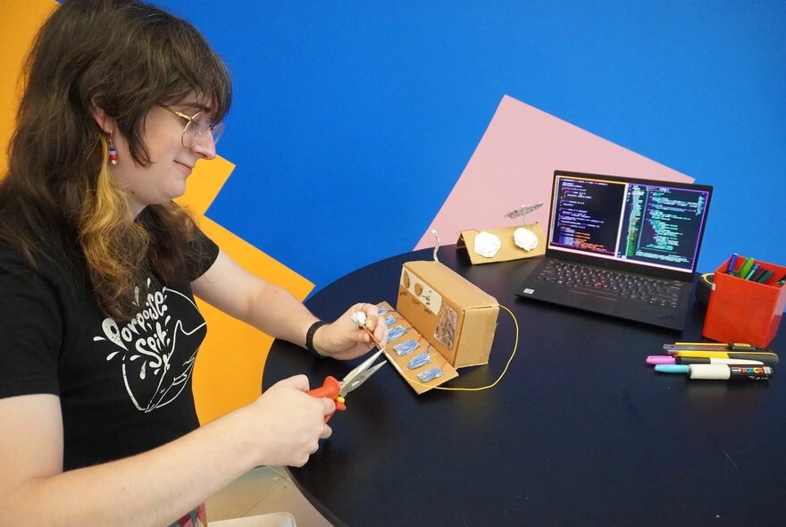 A person seated at a table using scissors to cut into a piece of cardboard, to make a simple sound controller, with a laptop and pens in the background.