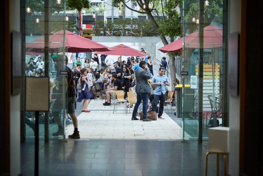 Looking through a building with glass walls and doors to a courtyard with red umbrellas and people gathered.
