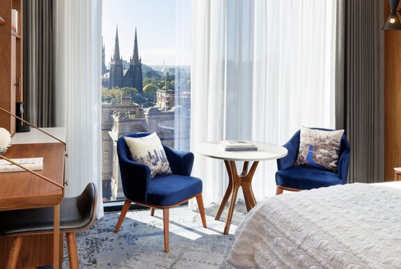 A hotel room showing the corner of a white-covered bed, two blue chairs and a small round table in front of a window with a view over Melbourne.
