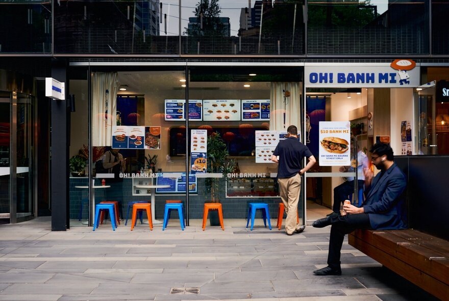 People outside a small takeaway Vietnamese banh mi shop with outdoor seating.