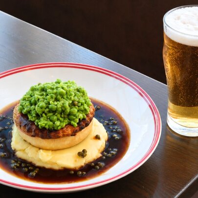 A pie resting on a bed of mashed potato surrounded by a pepper jus, a pint of beer on the table next to it.