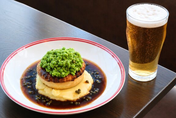 A pie resting on a bed of mashed potato surrounded by a pepper jus, a pint of beer on the table next to it.
