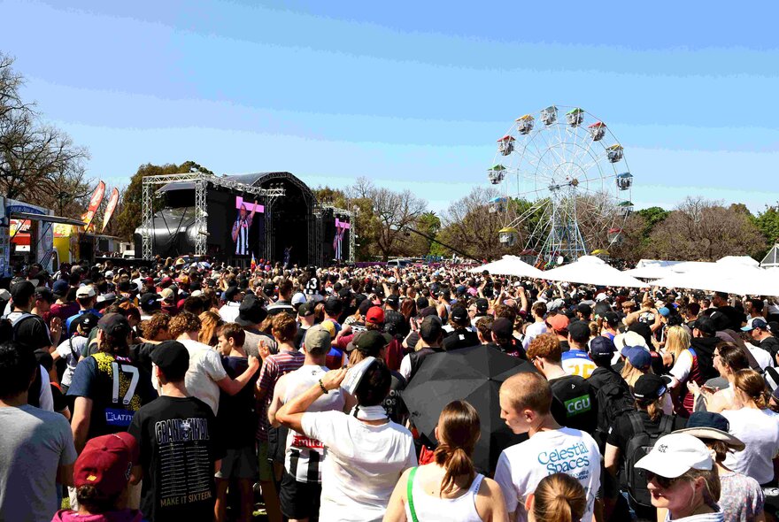 A crowd of people watching something on an outdoor stage, a ferris wheel in the background.