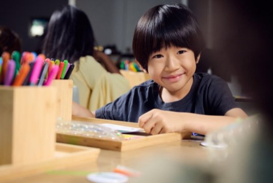 A smiling young boy, at a craft table engaged in an activity.