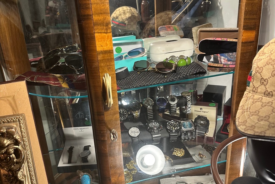 An antique wooden display cabinet, featuring glass doors and shelves filled with various collectible items and small accessories like watches and glasses.