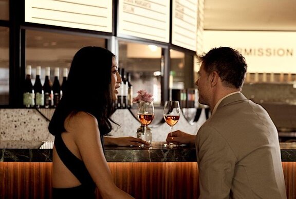 A young couple enjoying a glass of wine each, seated at a bar that looks like a cinema lobby.