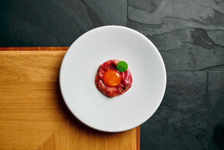 A white plate balanced on the corner of a wooden table with a small circle of sliced beef and a raw egg yolk on top. 