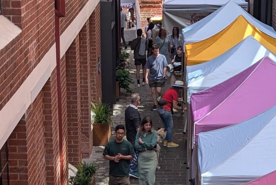 Tented market stalls lining an alley next to a brick building, with people browsing.