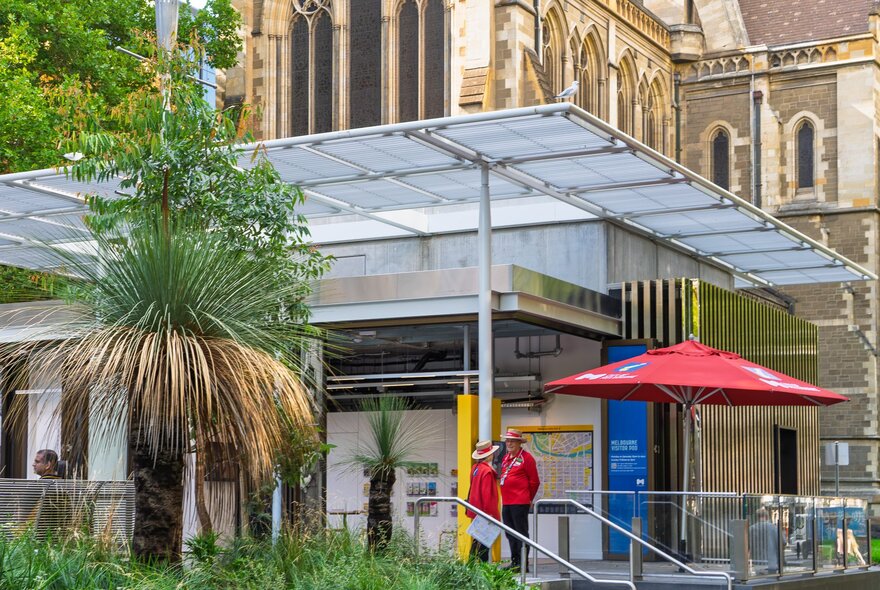 The Melbourne Visitor Pod in City Square, with a translucent roof and a large grass tree in the foreground, with two people in red jackets standing near the entrance with a red umbrella.
