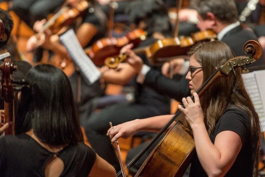 A cellist playing her cello amongst a full orchestra, seen out of focus in the background.