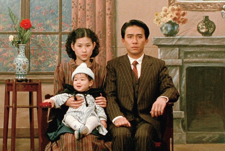 A still image from a Taiwanese film of a man, woman and baby sitting formally for a picture in a room with an illustrated formal background. 