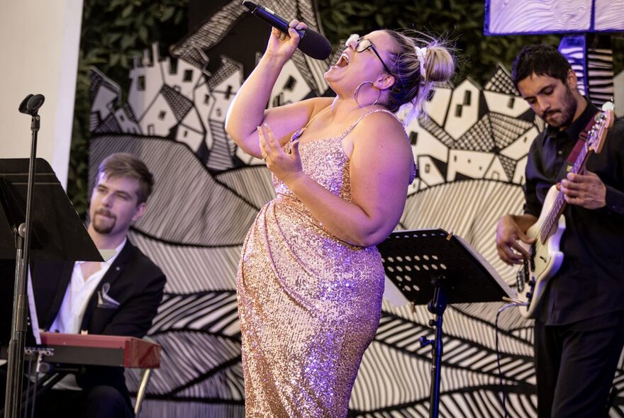 Performer Elly Poletti on stage in a shimmering sequined dress, singing into a microphone, with an electric guitarist and keyboard player behind her.