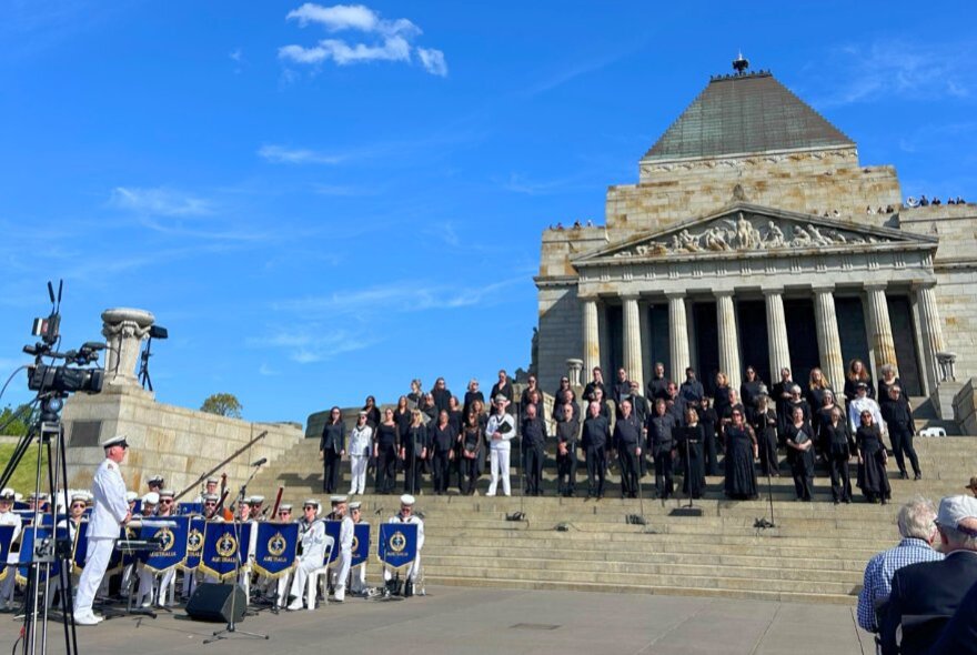 A chorus of singers standing on the steps of the Shrine of Remembrance, with the Royal Australian Navy Band playing their instruments on the forecourt, the Shrine building in the background, set against a blue sky.