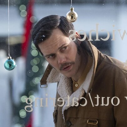 Still from a film showing a man with a mustache and a brown jacket looking through a shop window.