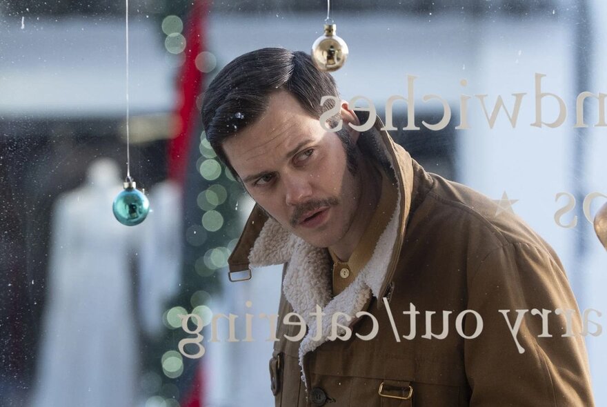 Still from a film showing a man with a mustache and a brown jacket looking through a shop window.