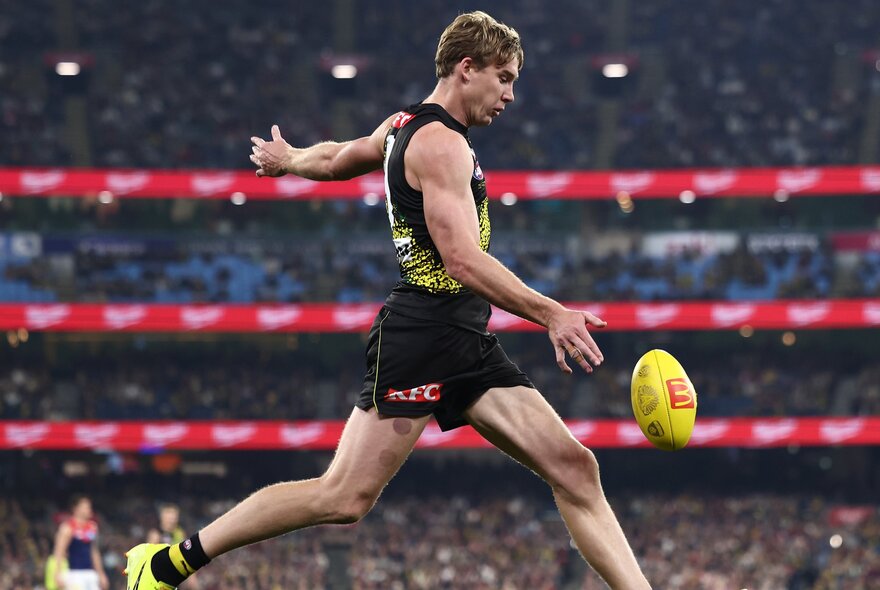 A Richmond AFL football player running and just about to kick the football on the field during a match, a blurred crowd of stadium fans in the background.