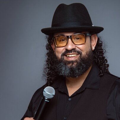 A man with a beard and curly hair, wearing a black fedora-style hat, yellow-tinted glasses, and a black shirt, holding a microphone and smiling at the camera. 