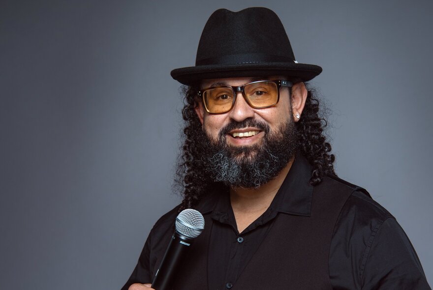 A man with a beard and curly hair, wearing a black fedora-style hat, yellow-tinted glasses, and a black shirt, holding a microphone and smiling at the camera. 