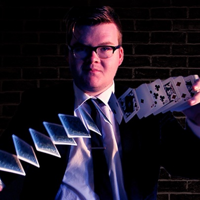 Magician Josh Staley wearing glasses, a dark suit and tie, shuffling a deck of cards.