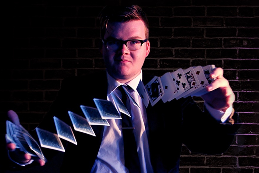 Magician Josh Staley wearing glasses, a dark suit and tie, shuffling a deck of cards.