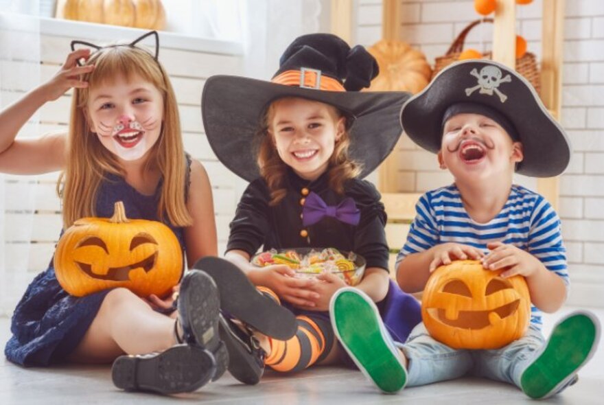 Three children seated on floor holding carved pumpkins, wearing Halloween outfits.