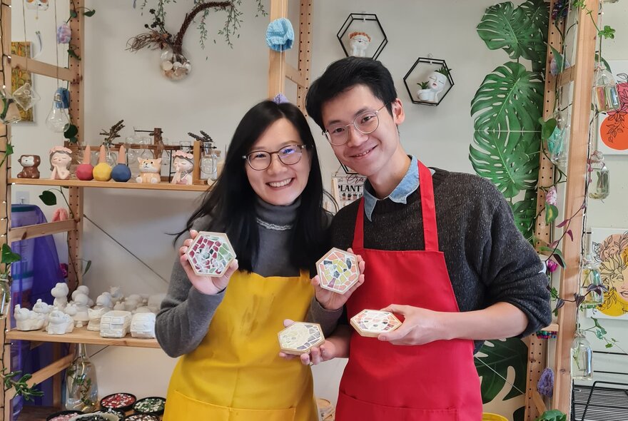 Workshop class participants smiling in a studio setting, holding up mosaic coasters they have made.
