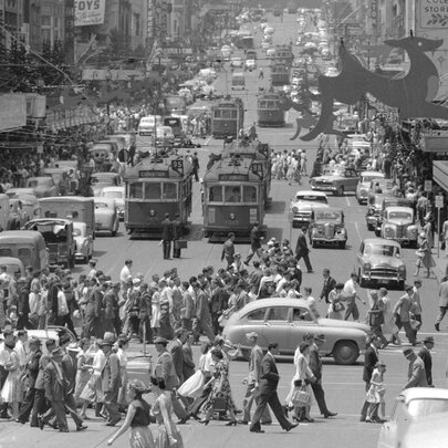 Black and white photograph of a busy Melbourne city intersection with trams, cars, people crossing the road, and buildings, dating from around the 1950s or 1960s.