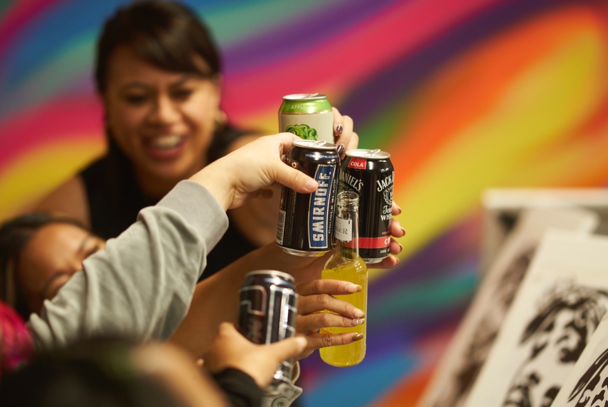 Hands holding up cans and bottles of drink in a toast, with canvases on easels in the foreground, as part of a sip and paint workshop.