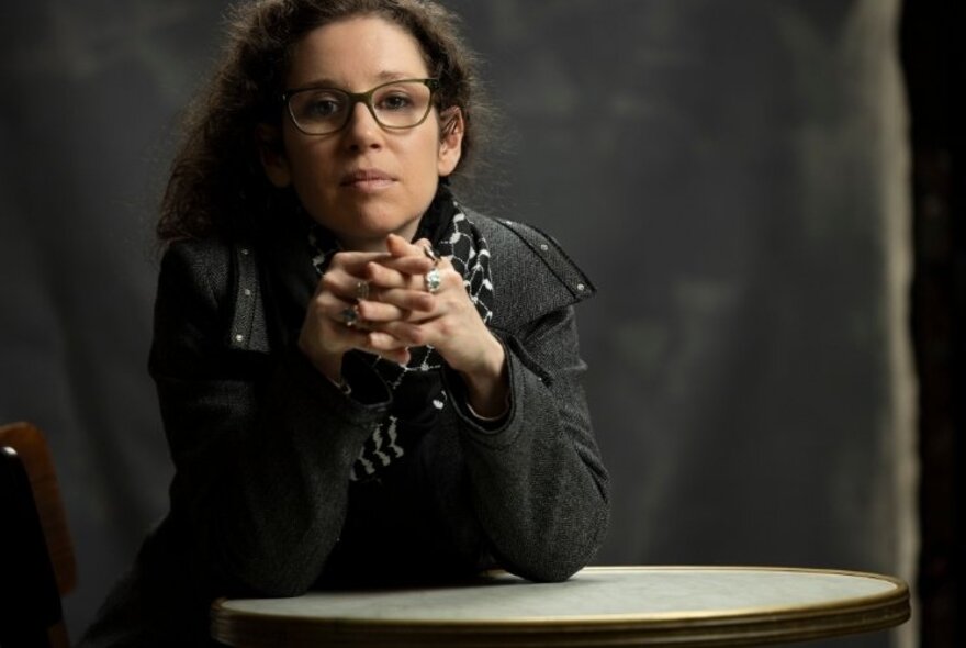 Micaela Sahhar, a woman seated at a small table, wearing a leather jacket, glasses and with long curly hair and her hands clasped together in front of her, elbows resting on the table.