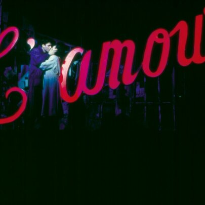 A couple on stage kissing in front of a red cursive sign saying L'amour.