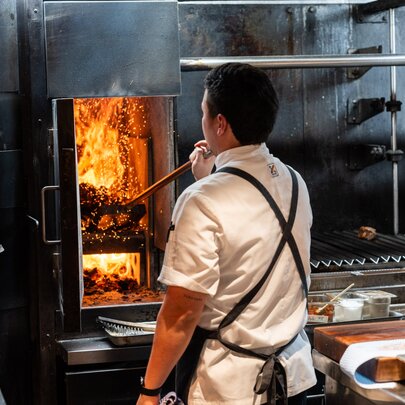 The back of a chef wearing an apron stoking a wood fired oven in a kitchen