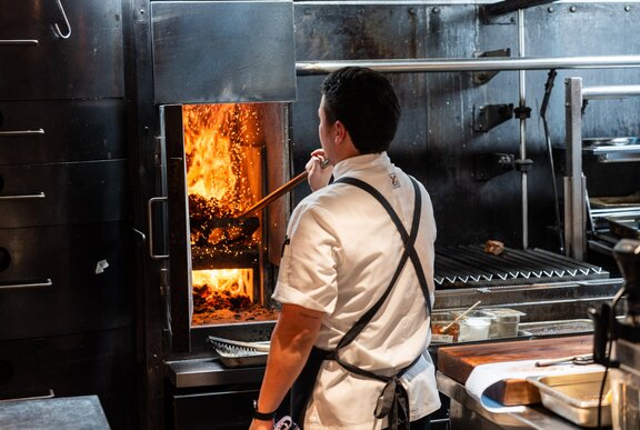 The back of a chef wearing an apron stoking a wood fired oven in a kitchen