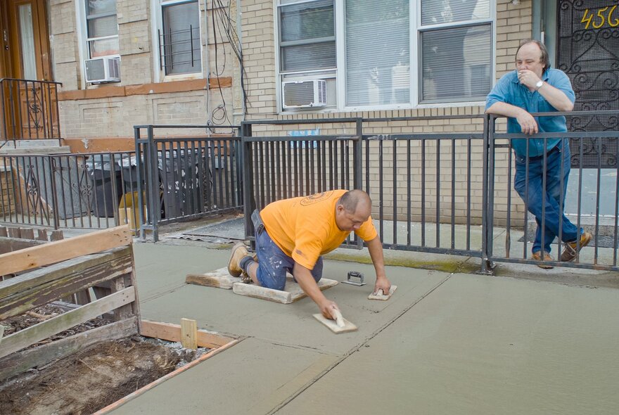 A still from the documentary film, The History of Concrete, shows a worker wearing an orange shirt kneeling on a footpath smoothing wet concrete, while another man in a blue shirt leans against a fence watching him.