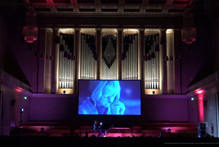 A solo pianist playing a grand piano on a stage in a large theatre, with a video screen projection from the game Final Fantasy behind them.