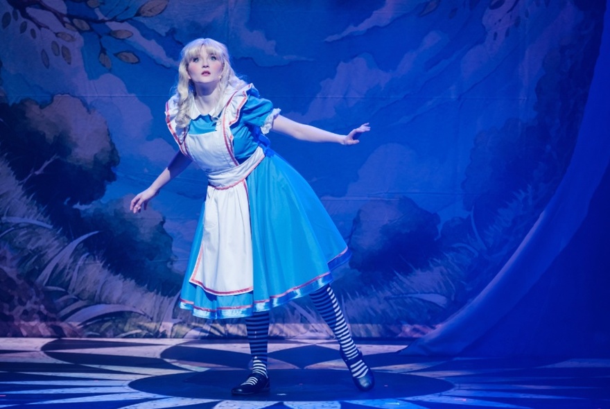 Actor wearing Alice in Wonderland blue dress and apron, standing with arms open looking out from the stage.