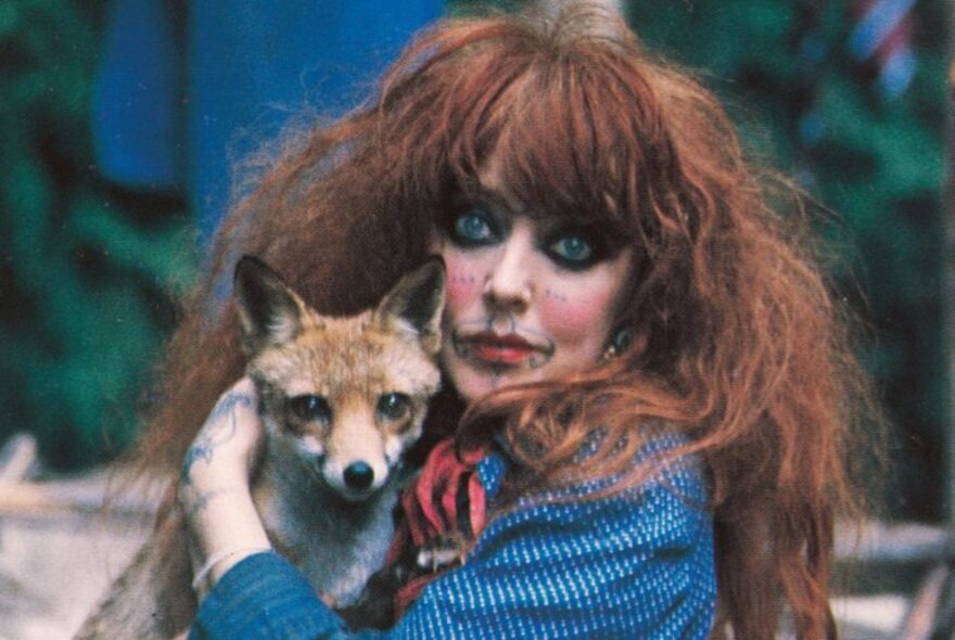 Portrait of Australian artist and dancer, Vali Myers, with red hair, thick black eye makeup and tattoos on her face and hands, holding her pet fox.
