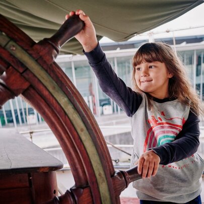 A young girl holds an old ship steering wheel.